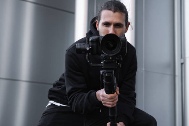 istock Young Professional videographer holding professional camera on 3-axis gimbal stabilizer. Pro equipment helps to make high quality video without shaking. Cameraman wearing white hoodie making a videos. 1216105766