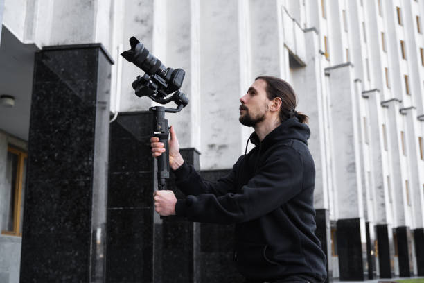 istock Young Professional videographer holding professional camera on 3-axis gimbal stabilizer. Pro equipment helps to make high quality video without shaking. Cameraman wearing black hoodie making a videos. 1216106389