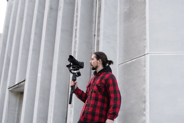 istock Young Professional videographer holding professional camera on 3-axis gimbal stabilizer. Pro equipment helps to make high quality video without shaking. Cameraman wearing red shirt making a videos. 1216106452