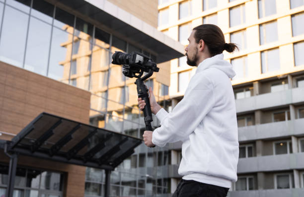 istock Young Professional videographer holding professional camera on 3-axis gimbal stabilizer. Pro equipment helps to make high quality video without shaking. Cameraman wearing white hoodie making a videos. 1216106503