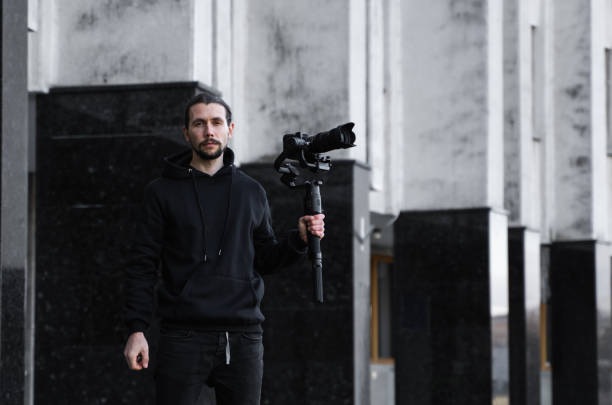 istock Young Professional videographer holding professional camera on 3-axis gimbal stabilizer. Pro equipment helps to make high quality video without shaking. Cameraman wearing black hoodie making a videos. 1216106626