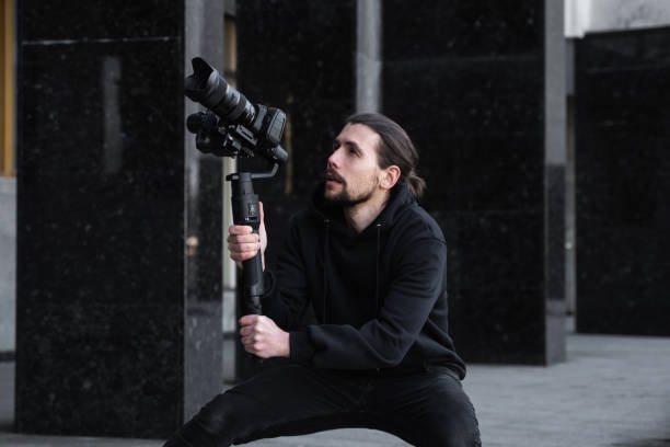 istock Young Professional videographer holding professional camera on 3-axis gimbal stabilizer. Pro equipment helps to make high quality video without shaking. Cameraman wearing black hoodie making a videos. 1216106679