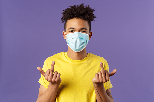 Medicine, covid19, coronavirus and people concept. Fuck you virus, unbothered, smart young hipster guy in medical mask show middle fingers and smile with eyes, staying protected indoors.