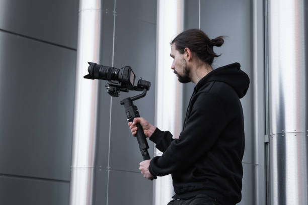 istock Young Professional videographer holding professional camera on 3-axis gimbal stabilizer. Pro equipment helps to make high quality video without shaking. Cameraman wearing white hoodie making a videos. 1216106892