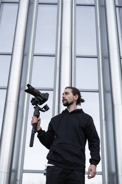istock Young Professional videographer holding professional camera on 3-axis gimbal stabilizer. Pro equipment helps to make high quality video without shaking. Cameraman wearing white hoodie making a videos. 1216107001