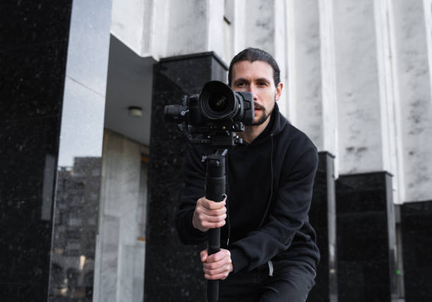 istock Young Professional videographer holding professional camera on 3-axis gimbal stabilizer. Pro equipment helps to make high quality video without shaking. Cameraman wearing black hoodie making a videos. 1216107741