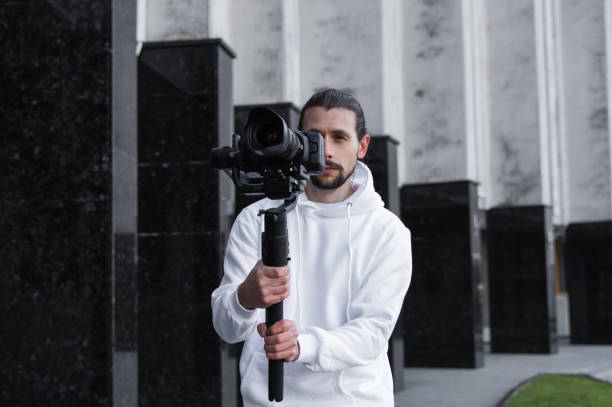 istock Young Professional videographer holding professional camera on 3-axis gimbal stabilizer. Pro equipment helps to make high quality video without shaking. Cameraman wearing white hoodie making a videos. 1216107978