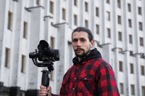 istock Young Professional videographer holding professional camera on 3-axis gimbal stabilizer. Pro equipment helps to make high quality video without shaking. Cameraman wearing red shirt making a videos. 1216108262