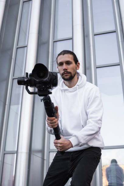 istock Young Professional videographer holding professional camera on 3-axis gimbal stabilizer. Pro equipment helps to make high quality video without shaking. Cameraman wearing white hoodie making a videos. 1216108415
