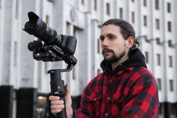 istock Young Professional videographer holding professional camera on 3-axis gimbal stabilizer. Pro equipment helps to make high quality video without shaking. Cameraman wearing red shirt making a videos. 1216108480