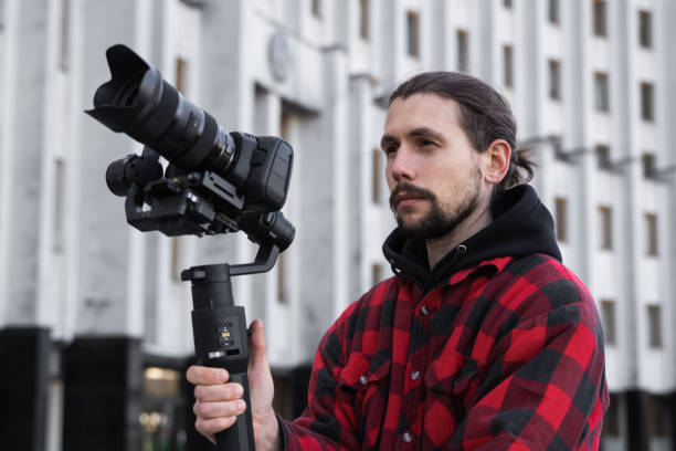 Young Professional videographer holding professional camera on 3-axis gimbal stabilizer. Pro equipment helps to make high quality video without shaking. Cameraman wearing red shirt making a videos. stock photo