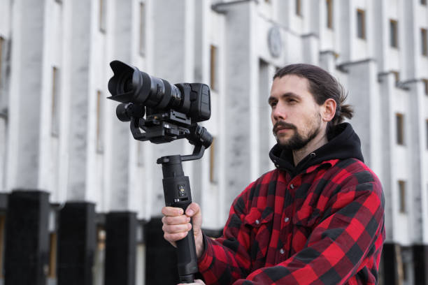 istock Young Professional videographer holding professional camera on 3-axis gimbal stabilizer. Pro equipment helps to make high quality video without shaking. Cameraman wearing red shirt making a videos. 1216108645