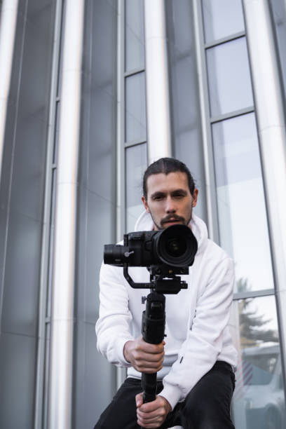 istock Young Professional videographer holding professional camera on 3-axis gimbal stabilizer. Pro equipment helps to make high quality video without shaking. Cameraman wearing white hoodie making a videos. 1216109439