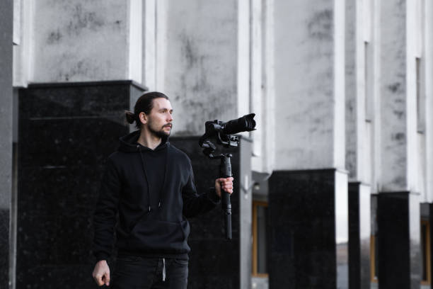 istock Young Professional videographer holding professional camera on 3-axis gimbal stabilizer. Pro equipment helps to make high quality video without shaking. Cameraman wearing black hoodie making a videos. 1216109553