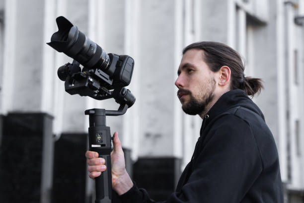 istock Young Professional videographer holding professional camera on 3-axis gimbal stabilizer. Pro equipment helps to make high quality video without shaking. Cameraman wearing black hoodie making a videos. 1216109642