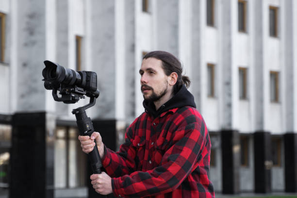 istock Young Professional videographer holding professional camera on 3-axis gimbal stabilizer. Pro equipment helps to make high quality video without shaking. Cameraman wearing red shirt making a videos. 1216112455