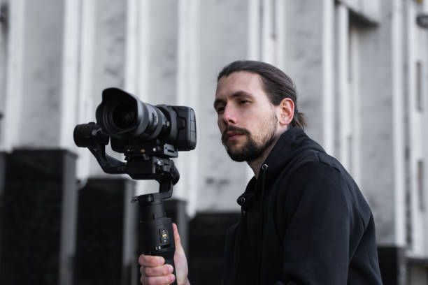istock Young Professional videographer holding professional camera on 3-axis gimbal stabilizer. Pro equipment helps to make high quality video without shaking. Cameraman wearing black hoodie making a videos. 1216112621