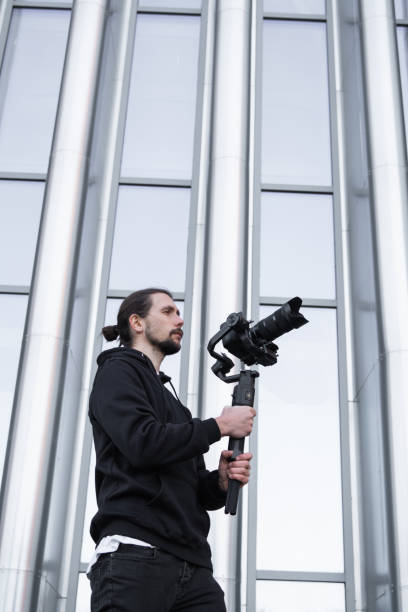 istock Young Professional videographer holding professional camera on 3-axis gimbal stabilizer. Pro equipment helps to make high quality video without shaking. Cameraman wearing white hoodie making a videos. 1216113100
