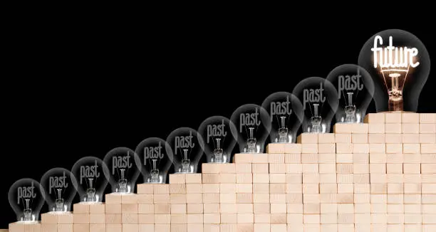 Large group of shining and dimmed light bulbs with fibers in a shape of Past and Future words on wooden block ladder isolated on black background. Concept of Innovation, Motivation and Success