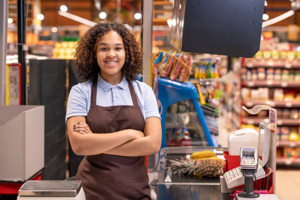 ziemlich junge lächelnde afroamerikanische verkäuferin, die sie anschaut - kassiererin supermarkt stock-fotos und bilder