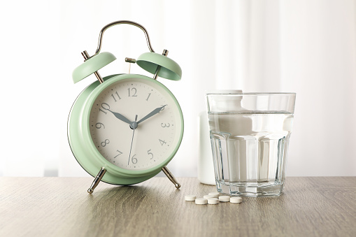Alarm clock, pills and glass of water on wooden table, close up