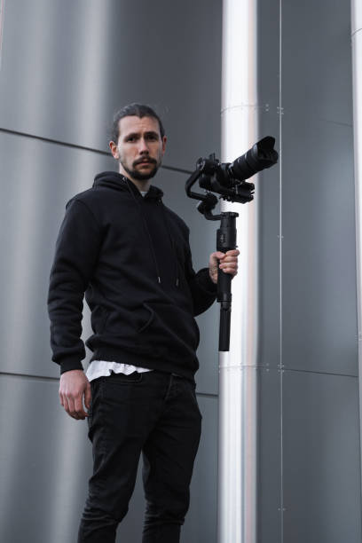 Young Professional videographer holding professional camera on 3-axis gimbal stabilizer. Pro equipment helps to make high quality video without shaking. Cameraman wearing white hoodie making a videos.