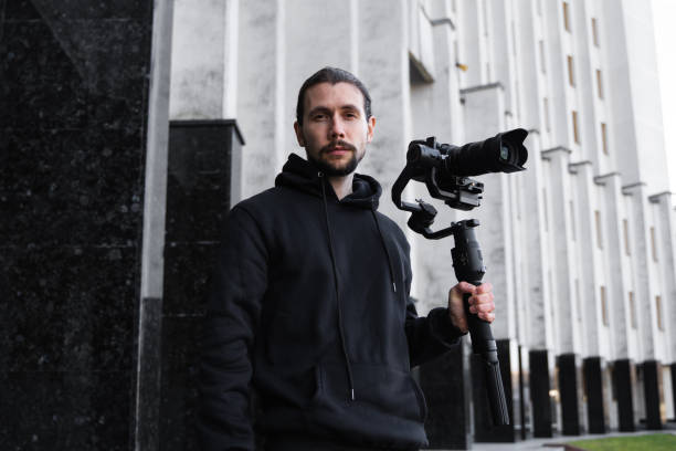 istock Young Professional videographer holding professional camera on 3-axis gimbal stabilizer. Pro equipment helps to make high quality video without shaking. Cameraman wearing black hoodie making a videos. 1216125349
