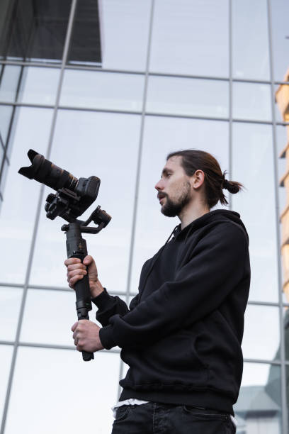 istock Young Professional videographer holding professional camera on 3-axis gimbal stabilizer. Pro equipment helps to make high quality video without shaking. Cameraman wearing white hoodie making a videos. 1216125554