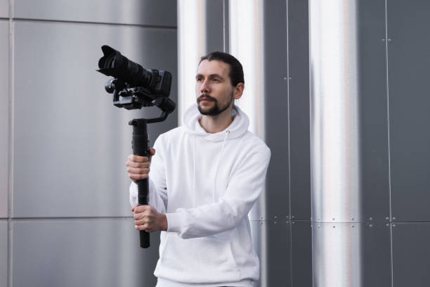 istock Young Professional videographer holding professional camera on 3-axis gimbal stabilizer. Pro equipment helps to make high quality video without shaking. Cameraman wearing white hoodie making a videos. 1216126292