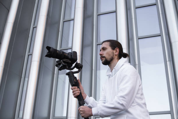 istock Young Professional videographer holding professional camera on 3-axis gimbal stabilizer. Pro equipment helps to make high quality video without shaking. Cameraman wearing white hoodie making a videos. 1216126860