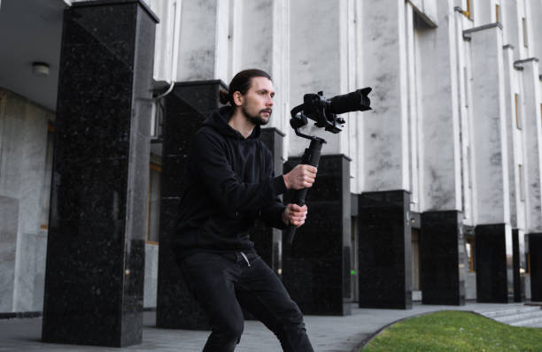 istock Young Professional videographer holding professional camera on 3-axis gimbal stabilizer. Pro equipment helps to make high quality video without shaking. Cameraman wearing black hoodie making a videos. 1216126902