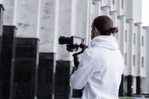 istock Young Professional videographer holding professional camera on 3-axis gimbal stabilizer. Pro equipment helps to make high quality video without shaking. Cameraman wearing white hoodie making a videos. 1216126981