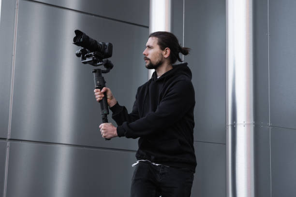 istock Young Professional videographer holding professional camera on 3-axis gimbal stabilizer. Pro equipment helps to make high quality video without shaking. Cameraman wearing white hoodie making a videos. 1216129054