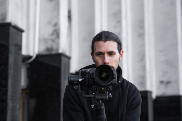 istock Young Professional videographer holding professional camera on 3-axis gimbal stabilizer. Pro equipment helps to make high quality video without shaking. Cameraman wearing black hoodie making a videos. 1216129129