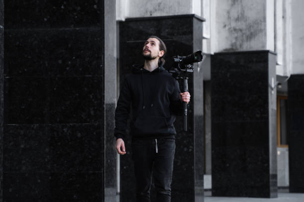istock Young Professional videographer holding professional camera on 3-axis gimbal stabilizer. Pro equipment helps to make high quality video without shaking. Cameraman wearing black hoodie making a videos. 1216129540