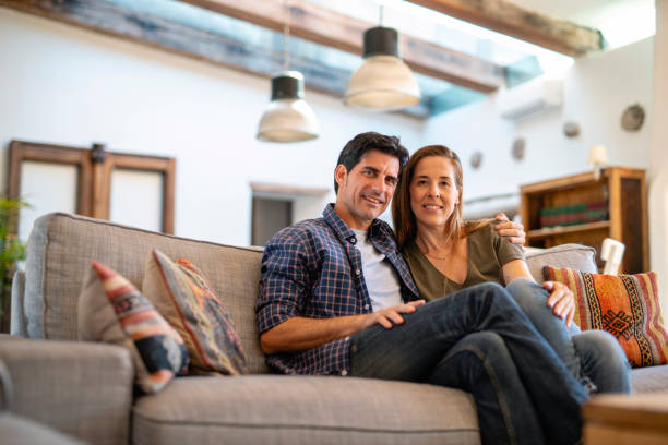 Relaxed Mature Couple Sitting Close on Sofa in Family Home Low angle close-up of Spanish couple in early 40s sitting close together on sofa in family home and smiling at camera. two adults stock pictures, royalty-free photos & images