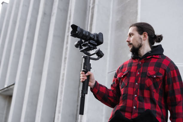 istock Young Professional videographer holding professional camera on 3-axis gimbal stabilizer. Pro equipment helps to make high quality video without shaking. Cameraman wearing red shirt making a videos. 1216130030