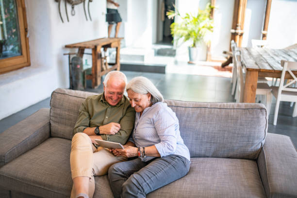 Senior Couple Sitting Together on Sofa with Digital Tablet Elevated view of smiling Spanish seniors in their 60s sitting side by side on sofa and using digital tablet in family home. farmhouse photos stock pictures, royalty-free photos & images