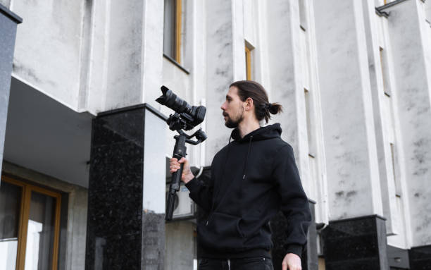 istock Young Professional videographer holding professional camera on 3-axis gimbal stabilizer. Pro equipment helps to make high quality video without shaking. Cameraman wearing black hoodie making a videos. 1216130250