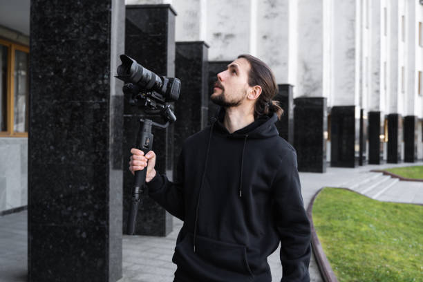 istock Young Professional videographer holding professional camera on 3-axis gimbal stabilizer. Pro equipment helps to make high quality video without shaking. Cameraman wearing black hoodie making a videos. 1216130825