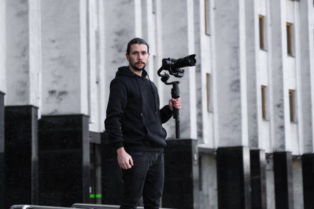 istock Young Professional videographer holding professional camera on 3-axis gimbal stabilizer. Pro equipment helps to make high quality video without shaking. Cameraman wearing black hoodie making a videos. 1216130907