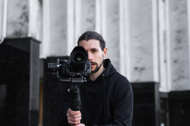 istock Young Professional videographer holding professional camera on 3-axis gimbal stabilizer. Pro equipment helps to make high quality video without shaking. Cameraman wearing black hoodie making a videos. 1216131099