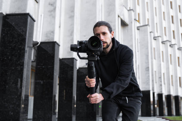 istock Young Professional videographer holding professional camera on 3-axis gimbal stabilizer. Pro equipment helps to make high quality video without shaking. Cameraman wearing black hoodie making a videos. 1216131234