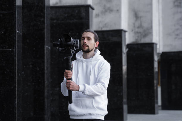 istock Young Professional videographer holding professional camera on 3-axis gimbal stabilizer. Pro equipment helps to make high quality video without shaking. Cameraman wearing white hoodie making a videos. 1216133455
