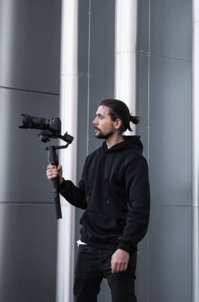 istock Young Professional videographer holding professional camera on 3-axis gimbal stabilizer. Pro equipment helps to make high quality video without shaking. Cameraman wearing white hoodie making a videos. 1216133557