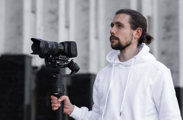 istock Young Professional videographer holding professional camera on 3-axis gimbal stabilizer. Pro equipment helps to make high quality video without shaking. Cameraman wearing white hoodie making a videos. 1216133612