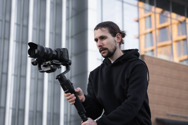 istock Young Professional videographer holding professional camera on 3-axis gimbal stabilizer. Pro equipment helps to make high quality video without shaking. Cameraman wearing white hoodie making a videos. 1216133859