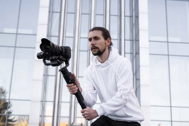 istock Young Professional videographer holding professional camera on 3-axis gimbal stabilizer. Pro equipment helps to make high quality video without shaking. Cameraman wearing white hoodie making a videos. 1216133915
