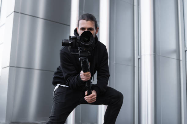 istock Young Professional videographer holding professional camera on 3-axis gimbal stabilizer. Pro equipment helps to make high quality video without shaking. Cameraman wearing white hoodie making a videos. 1216133961