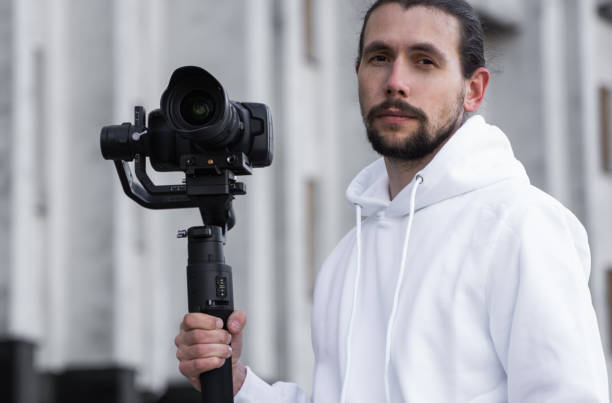 istock Young Professional videographer holding professional camera on 3-axis gimbal stabilizer. Pro equipment helps to make high quality video without shaking. Cameraman wearing white hoodie making a videos. 1216134016